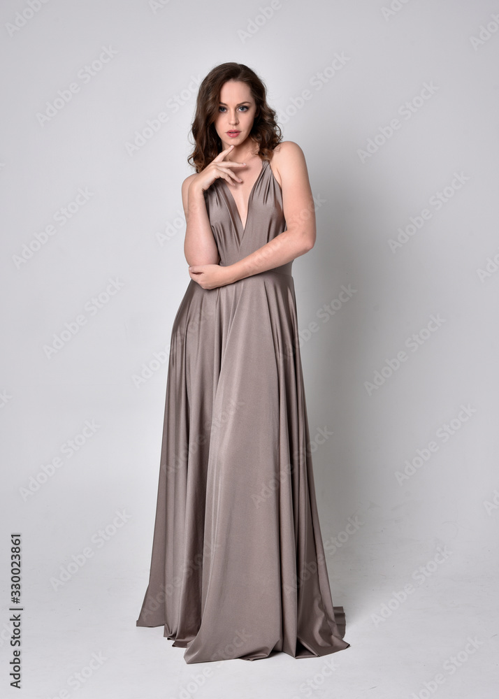  Portrait of a pretty brunette girl wearing a long silver evening gown, full length standing pose against a studio background.