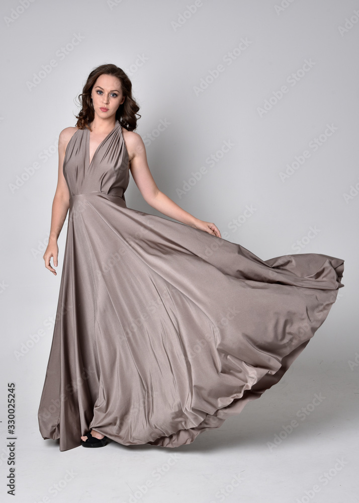 Portrait of a pretty brunette girl wearing a long silver evening gown, full length standing pose against a studio background. Portrait of a pretty brunette girl wearing a long silver evening gown, full length standing pose against a studio background.