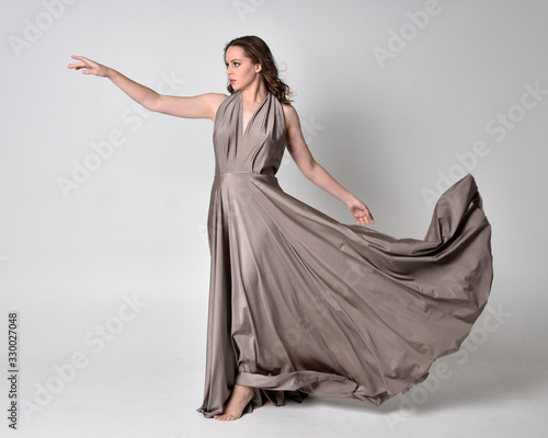  Portrait of a pretty brunette girl wearing a long silver evening gown, full length standing pose against a studio background.