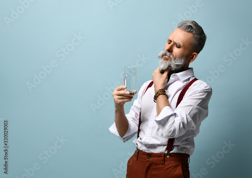Gray-haired aged man in white shirt, brown suspenders and bracelet. He is holding glass of water, posing against blue background