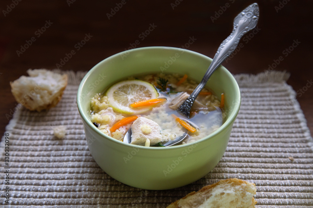 Delicious homemade chicken broth soup with lemon slice. Noodle soup in light green bowl and breadcrumbs on table napkin