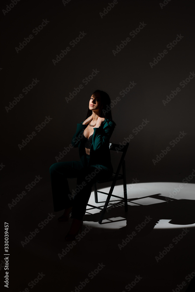 Obraz premium Studio photo of pretty brunette woman in twilight sitting on black chair. A ray of light hits her face.