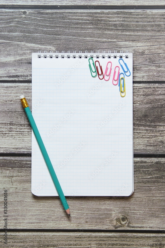 Notepad with a blank white sheet in a checker paper with simple graphite pencil and with paper clips lies on the background of wooden boards. Notepad with a blank white sheet in a checker paper with simple graphite pencil and with paper clips lies on the background of wooden boards.
