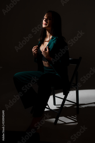 Studio photo of pretty brunette woman in twilight sitting on black chair. A ray of light hits her face.