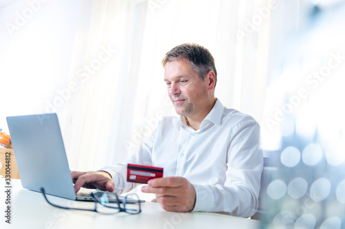 Caucasian middle aged smiling man  holding a red credit card and entering data into a laptop