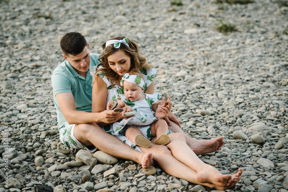 Family playing stones near water. Mom, dad hugging daughter sitting on beach near lake. The concept of summer holiday. Mother's, father's, baby's day. Spending time together. Family look. Family playing stones near water. Mom, dad hugging daughter sitting on beach near lake. The concept of summer holiday. Mother's, father's, baby's day. Spending time together. Family look.