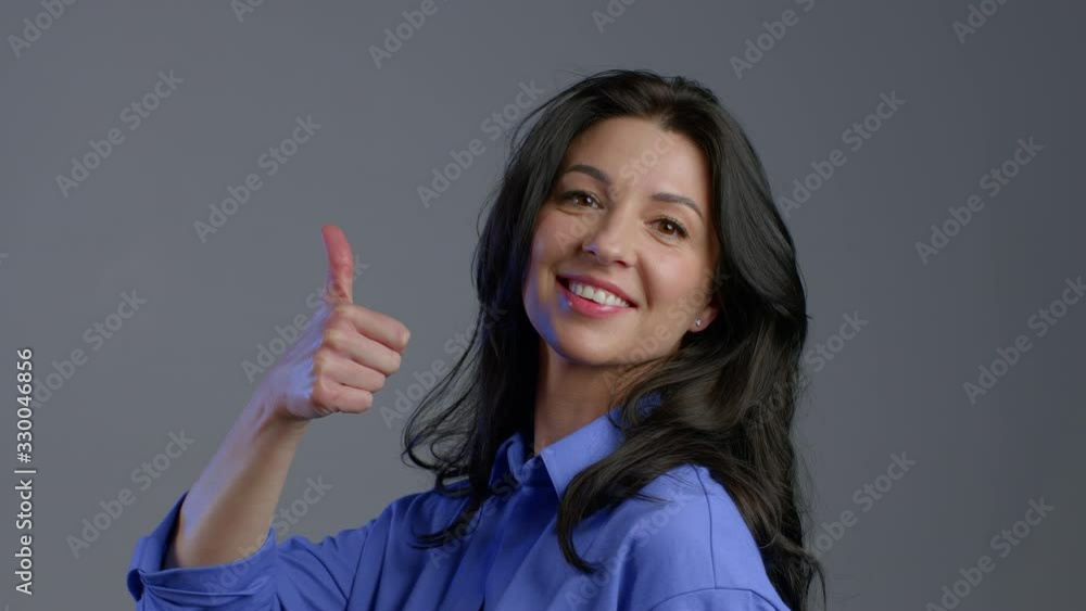 Happy adult woman on grey background. Lady with long hair in blue shirt shows yes like thumbs up gesture of victory,she achieved result, goals. Happy adult woman on grey background. Lady with long hair in blue shirt shows yes like thumbs up gesture of victory,she achieved result, goals.