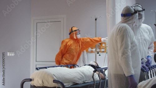 Doctor in Orange Protective Suit Turning On Coronavirus Pateint's Heart Monitor - Wide Dolly Shot photo