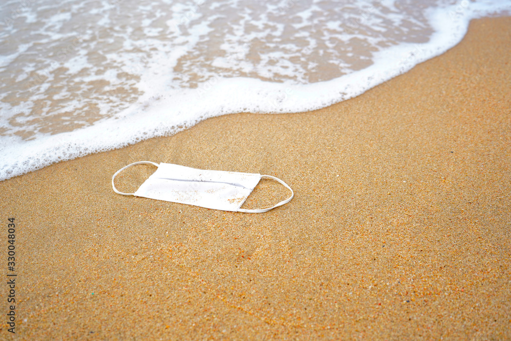 A white cotton face mask is left abandoned on the beach. It must be disposed in a proper hazardous waste bin, preventing germs spread. Objects and medical supply waste management. A white cotton face mask is left abandoned on the beach. It must be disposed in a proper hazardous waste bin, preventing germs spread. Objects and medical supply waste management.