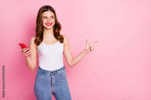 Portrait of positive cheerful girl use smart phone point index finger copyspace suggest select ads promotion wear white tank-top denim jeans isolated over pink color background