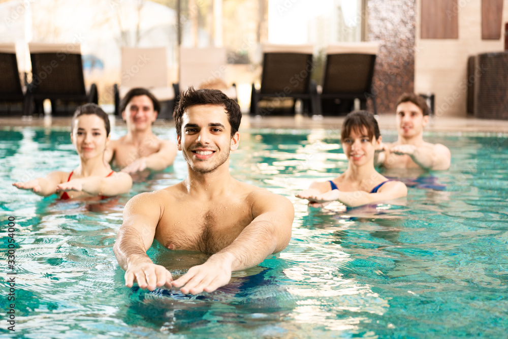 Selective focus of trainer smiling at camera while exercising water aerobics with people in swimming pool