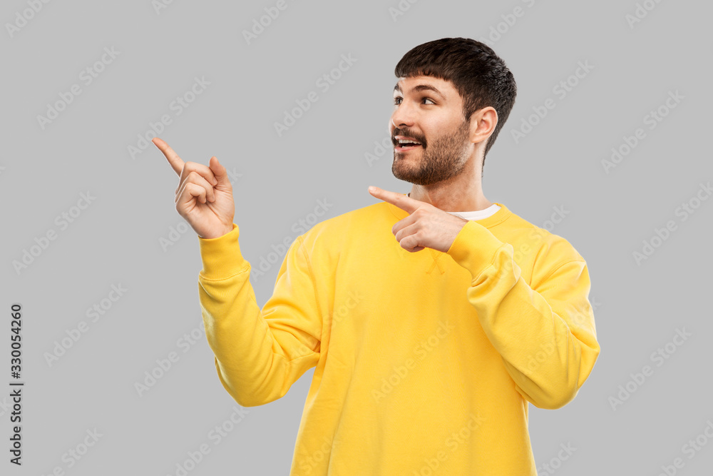 people concept - smiling young man in yellow sweatshirt pointing fingers to something over grey background people concept - smiling young man in yellow sweatshirt pointing fingers to something over grey background