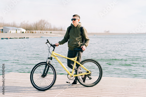 a young man in khaki sportswear and glasses stands on a pontoon near the lake and holds a yellow Bicycle near him on a Sunny spring day © Дарья Фомина