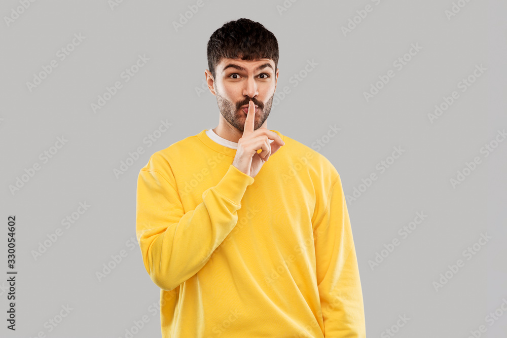 secret and people concept - smiling young man in yellow sweatshirt making hush gesture with finger on lips over grey background secret and people concept - smiling young man in yellow sweatshirt making hush gesture with finger on lips over grey background