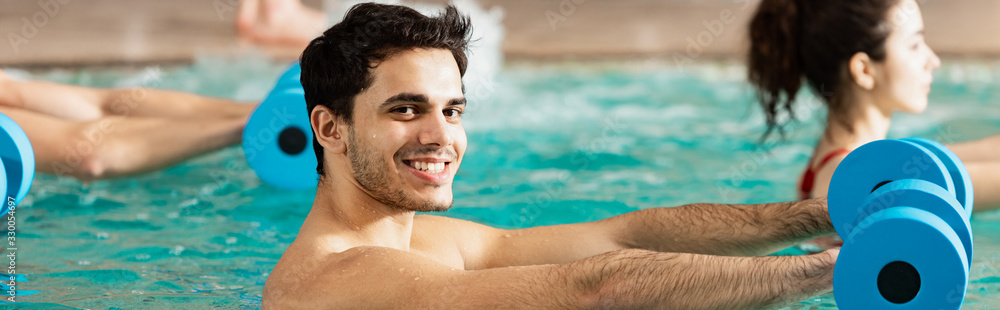 Side view of man smiling at camera while exercising with dumbbells in swimming pool, panoramic shot Side view of man smiling at camera while exercising with dumbbells in swimming pool, panoramic shot