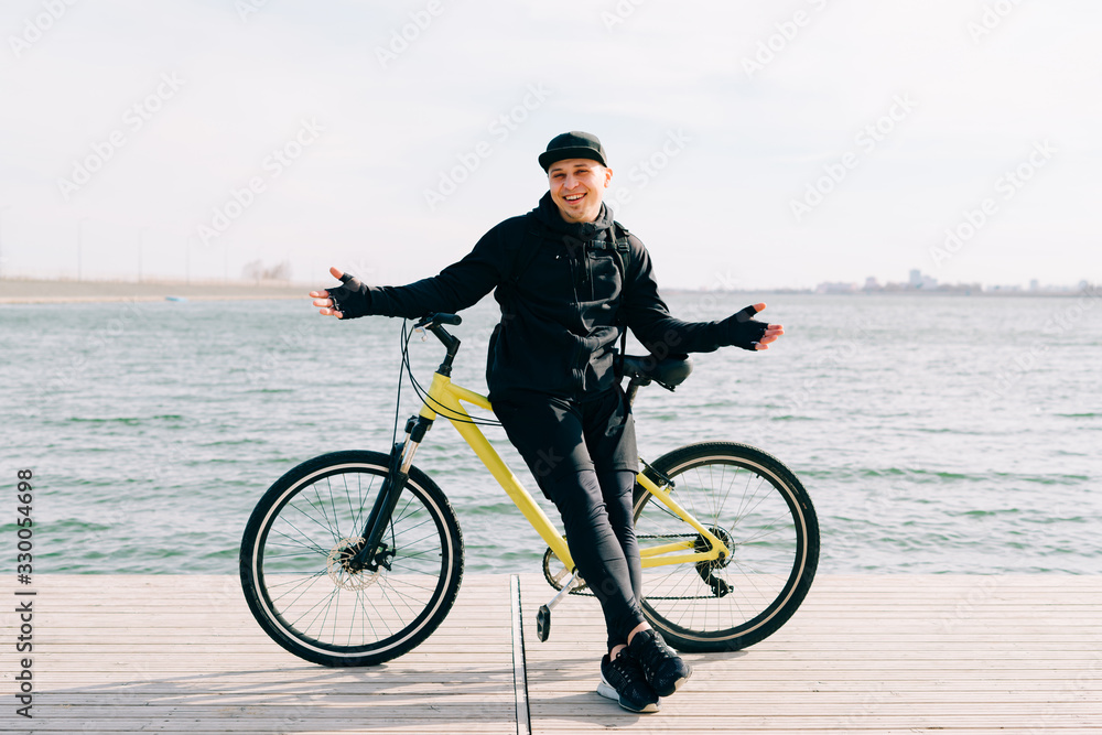 Fototapeta premium a young man in black sportswear stands on a pontoon near the lake and holds a yellow Bicycle near him on a Sunny spring day