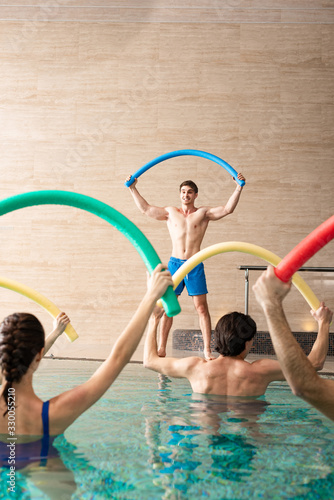 Fototapeta Naklejka Na Ścianę i Meble -  Smiling trainer with pool noodle exercising with group of young people in swimming pool