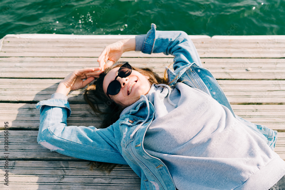 a young pretty girl in a denim jacket and glasses is lying on a pier near the lake on a Sunny spring day