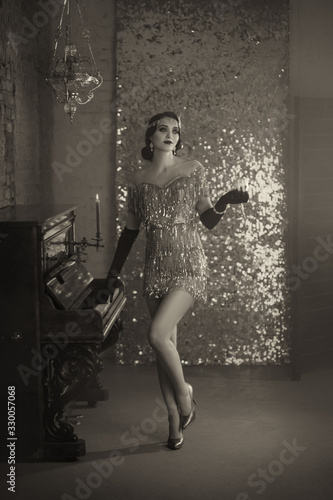 Black and white photo retro woman. finger wave hairstyle, short vintage silver shiny dress style 20s. Girl stands near old piano. backdrop romantic classic interior bright glitter wall, burning candle
