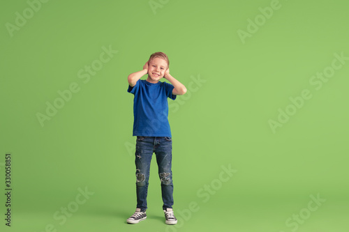 Covering ears. Happy boy playing and having fun on green studio background. Caucasian kid in bright cloth looks playful, laughting, smiling. Concept of education, childhood, emotions, facial © master1305