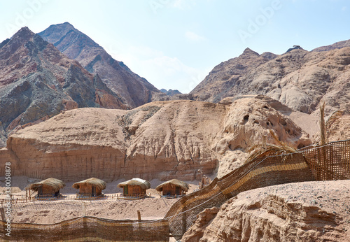 Canvas-taulu Sinai Peninsula, Egypt - Four traditional huts in a Bedouin village, nearby is a colored canyon, huts are thatched and fenced, behind huts are brown mountains