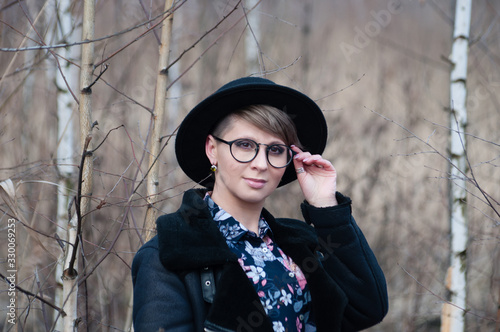 Pretty woman in glasses and a black hat posing among birches in early spring or late autumn