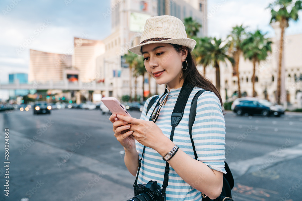 happy female travel backpacker with mobile phone while walking on city street. smiling girl tourist texting message on cellphone.  lady photographer standing on las vegas boulevard with coconut trees