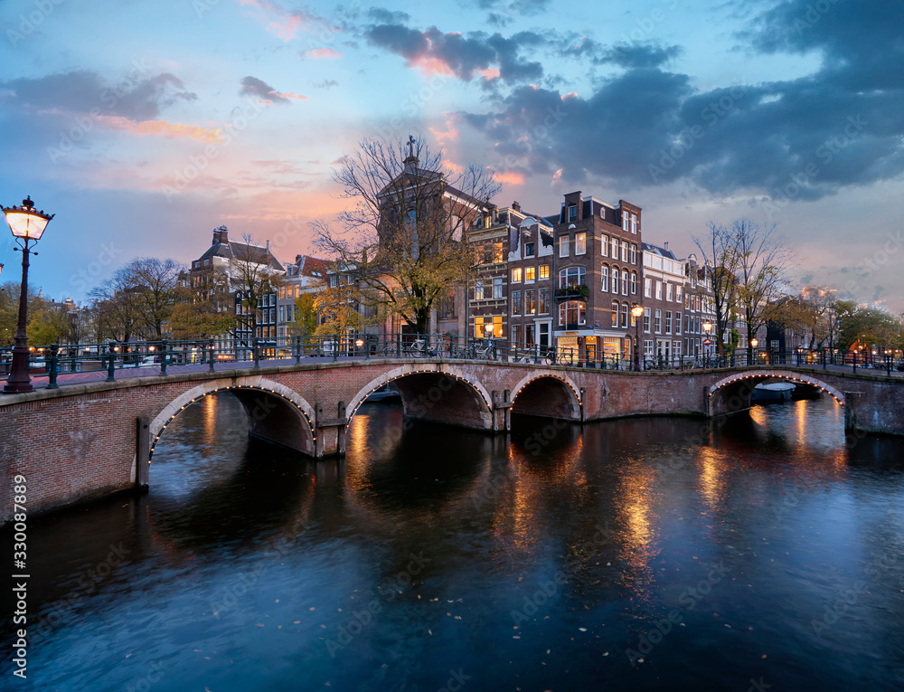Famous Amsterdam canals with historic houses and stone bridge just after sunset with colourful sky and bluehour lights                     