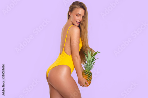 Sexy tanned long-haired brunette in yellow swimsuit posing with pineapple in her hands on white background