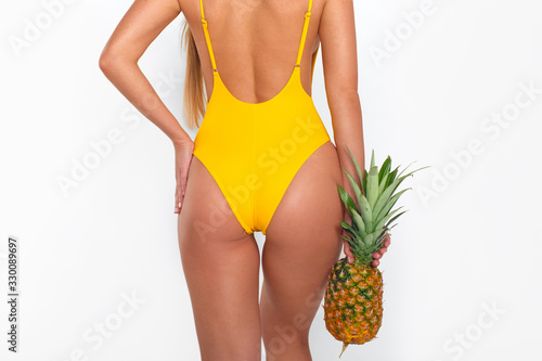Cropped picture of sexy tanned girl in yellow swimsuit posing with her back to camera with pineapple in her hands on white background