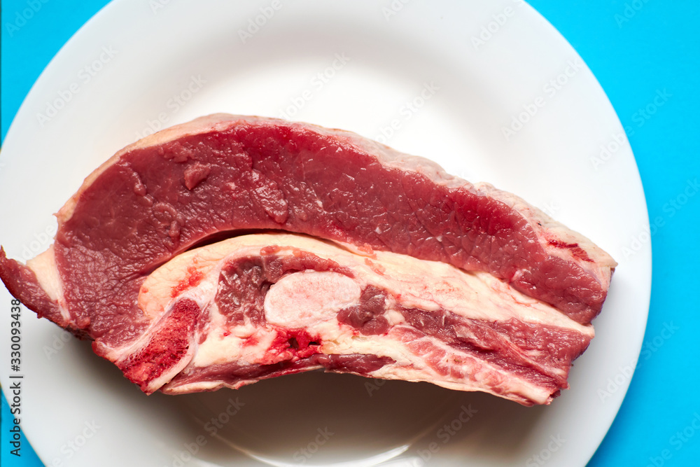 Raw veal steak on a white plate on a blue background. Raw veal steak on a white plate on a blue background.