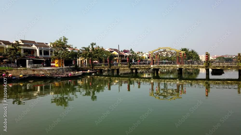 Hoi An, Quang Nam, Vietnam - March 9, 2020: Timelapse of people tourists walking on bridge over Thu Bon river, Hoi An, UNESCO World Heritage Site. Traditional vietnamese boats moored to shore. Sunny d Hoi An, Quang Nam, Vietnam - March 9, 2020: Timelapse of people tourists walking on bridge over Thu Bon river, Hoi An, UNESCO World Heritage Site. Traditional vietnamese boats moored to shore. Sunny d