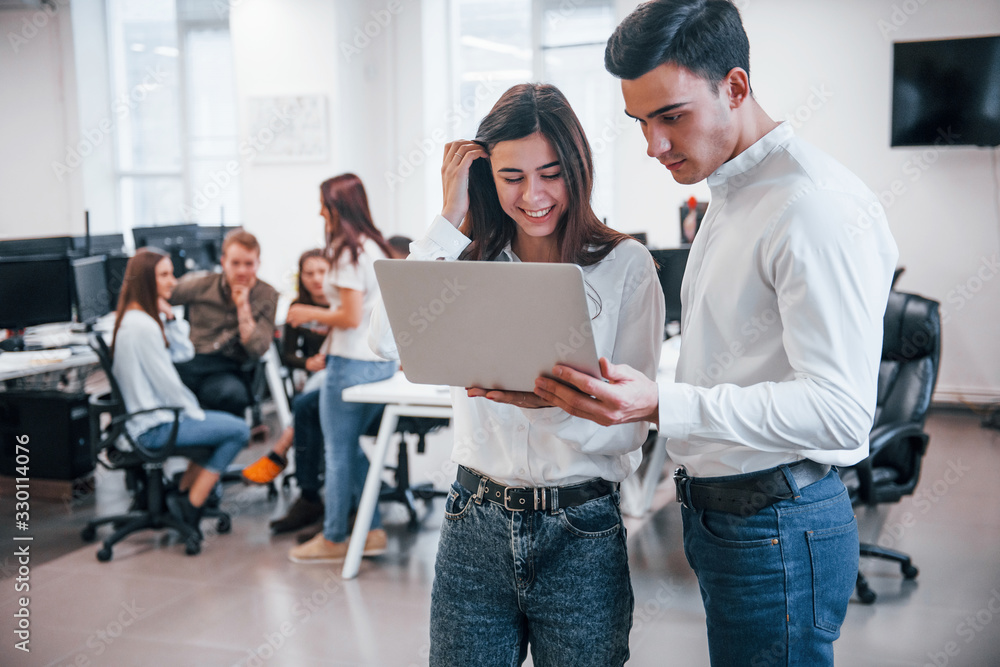 Man and woman in formal wear standing with laptop in hands indoors in office with young people that works behind them Man and woman in formal wear standing with laptop in hands indoors in office with young people that works behind them