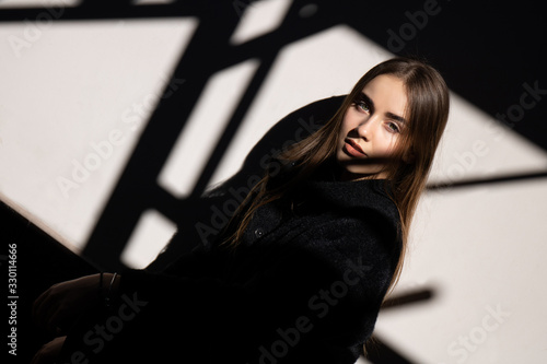 Portrait of young beautiful girl in hard light and shadow with long hair wearing a black coat on white background. Urban. Outdoor. Text space.