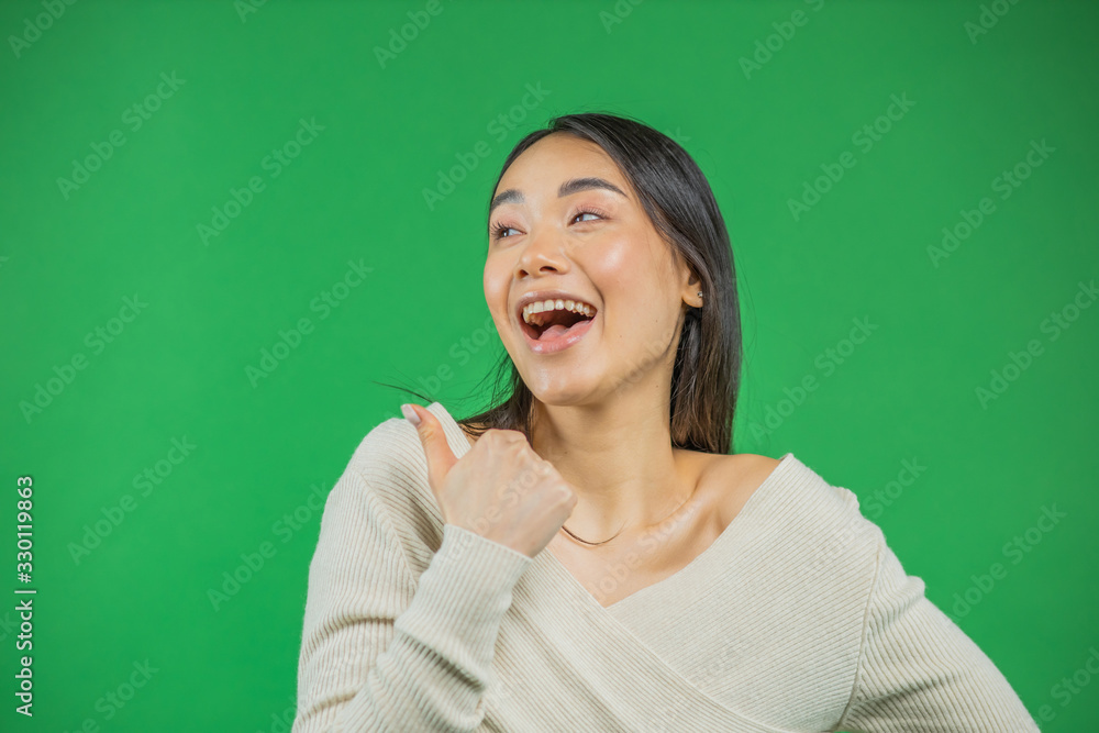 Portrait of an attractive young asian woman smiling looks away and showing thumbs up over green background Portrait of an attractive young asian woman smiling looks away and showing thumbs up over green background