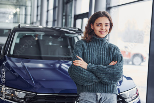 Cheerful young woman standing in front of modern new car indoors