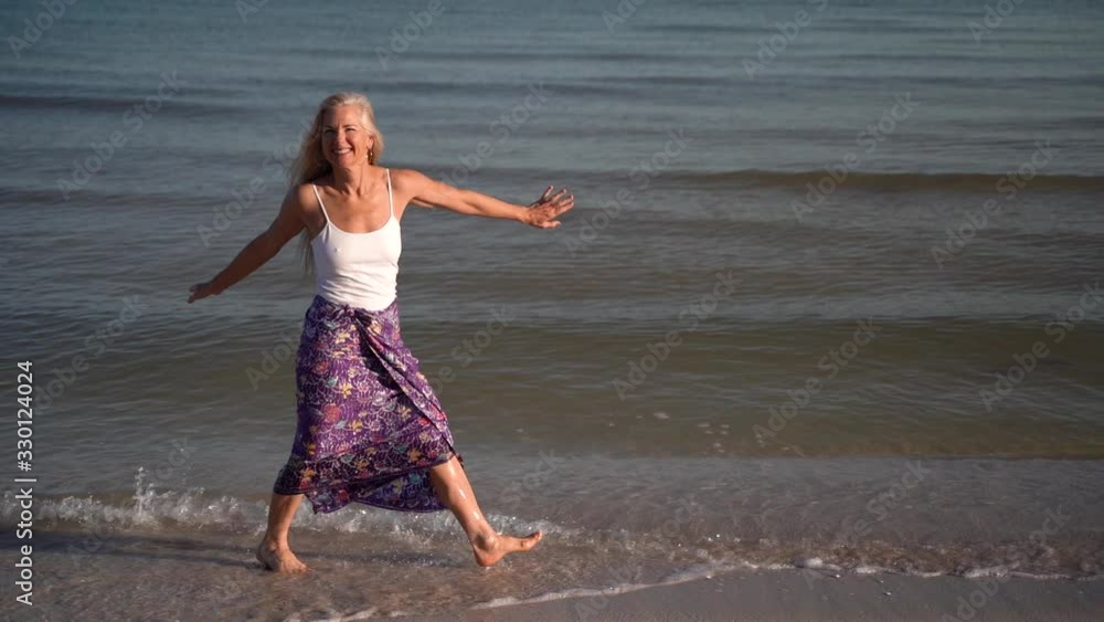 Slow motion of mature woman in sarong walking on a beach and splashing in the water at sunrise or sunset. Slow motion of mature woman in sarong walking on a beach and splashing in the water at sunrise or sunset.