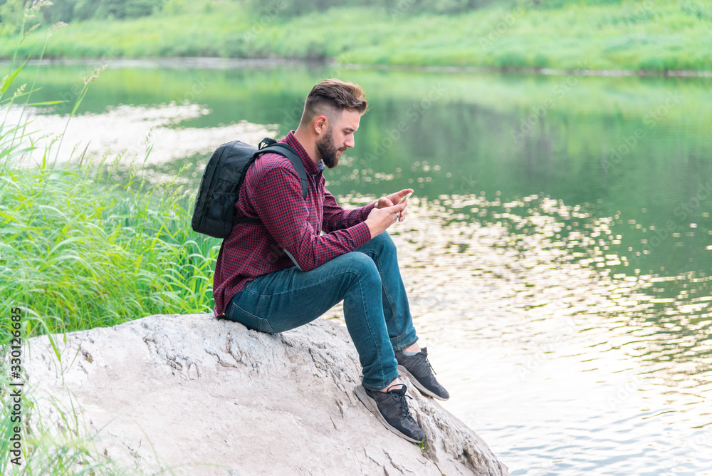 a man with a phone, with a backpack, in a shirt and jeans sits on a stone against the background of nature on a summer day. camping mobile concept a man with a phone, with a backpack, in a shirt and jeans sits on a stone against the background of nature on a summer day. camping mobile concept