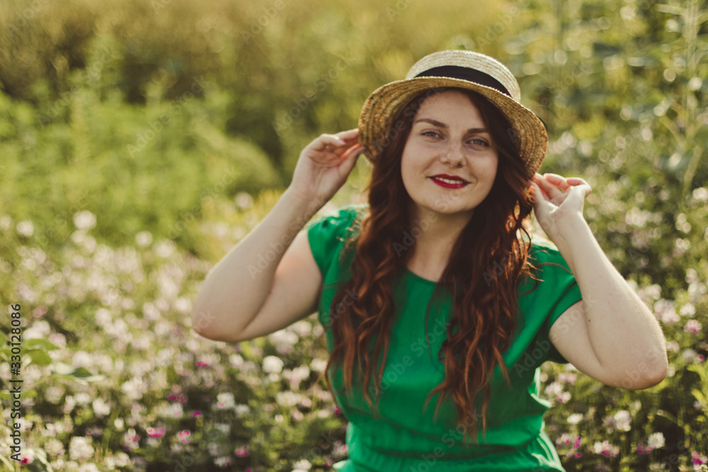 Young curly Caucasian girl in a green dress and a straw hat looks at the camera and smiles in a flower field on sunny day. Spring or summer panorama photo