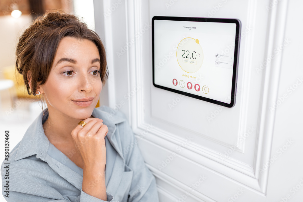 Young woman controlling temperature in the living room with a digital touch screen panel. Concept of heating control in a smart home Young woman controlling temperature in the living room with a digital touch screen panel. Concept of heating control in a smart home
