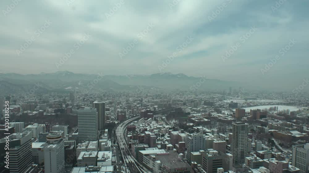 SAPPORO, HOKKAIDO, JAPAN - FEB 2020 : Aerial high angle view of cityscape of Sapporo city. View of railway, buildings and street traffic. Otaru area behind mountains. Time lapse shot in day time. SAPPORO, HOKKAIDO, JAPAN - FEB 2020 : Aerial high angle view of cityscape of Sapporo city. View of railway, buildings and street traffic. Otaru area behind mountains. Time lapse shot in day time.