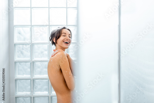 Beautiful naked woman taking a shower at home, standing under the water drops in the modern shower made of glass blocks