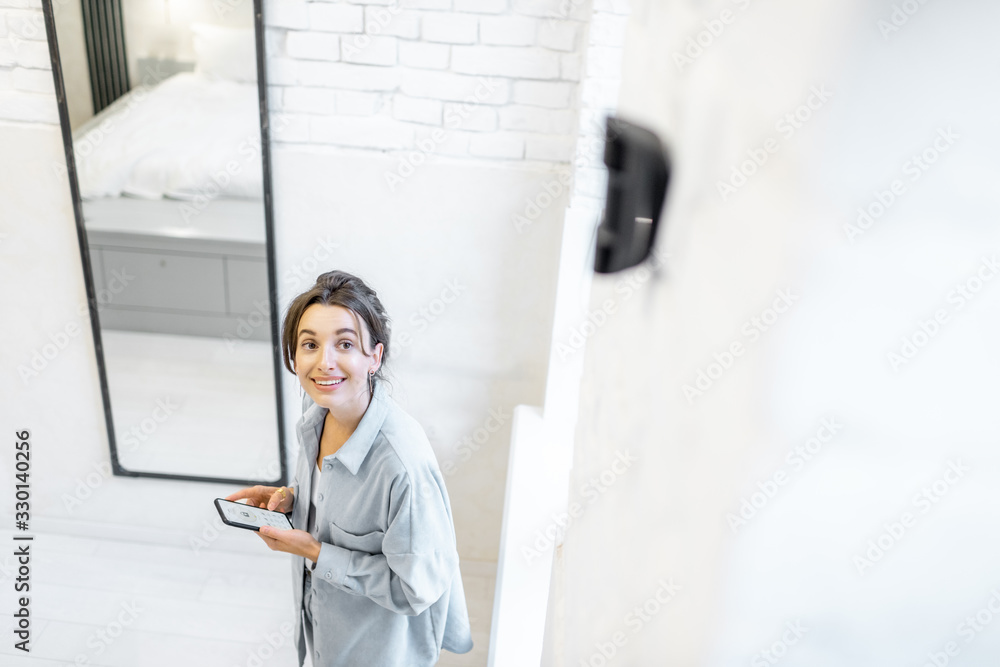 Woman controlling alarm system with a smart phone wireless, standing in the room with motion sensor mounted on the wall