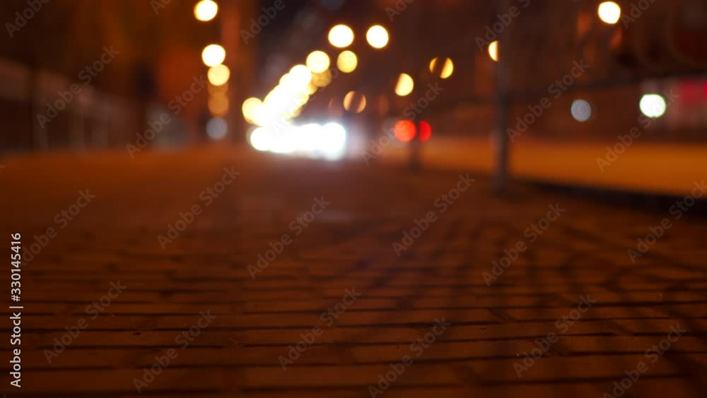 Night city street lights, surface level of dark stone tile sidewalk pavement and cars on the road.