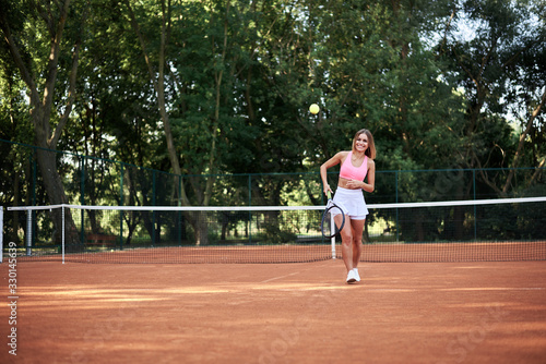 Young blond woman, wearing pink top and white skirt and sneakers, playing tennis on court in summer. Full-length portrait of sportswoman, training outside with tennis racket and light green ball. © Natalia