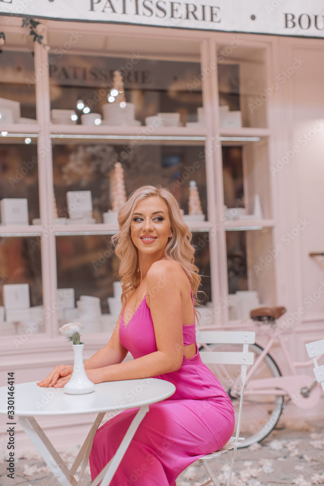 Obraz premium Attractive happy blonde in pink sexy dress sitting at the round cafe table in locations stylized as Paris confectionery