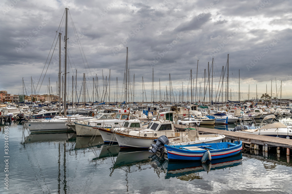Marina with docked yachts, sailboats and fishing boats on Ortygia island at sunrise with cloudy sky, province of Syracuse in Sicily