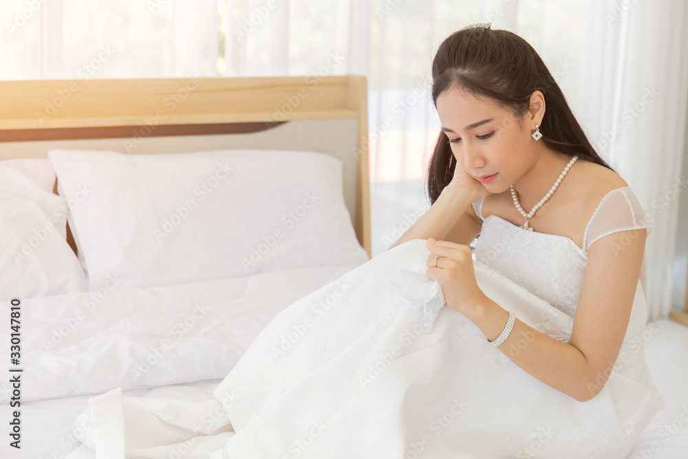 An Asian bride in a white wedding dress kneels down on the bed in her handkerchief to absorb her tears.