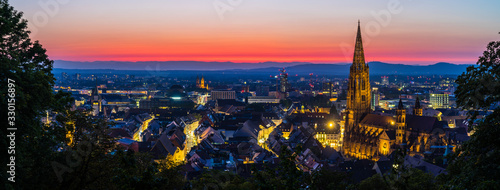Germany, XXL panorama of colorful red sunset sky decorating illuminated skyline of black forest city freiburg im breisgau, seen from above aerial after sunset in summer