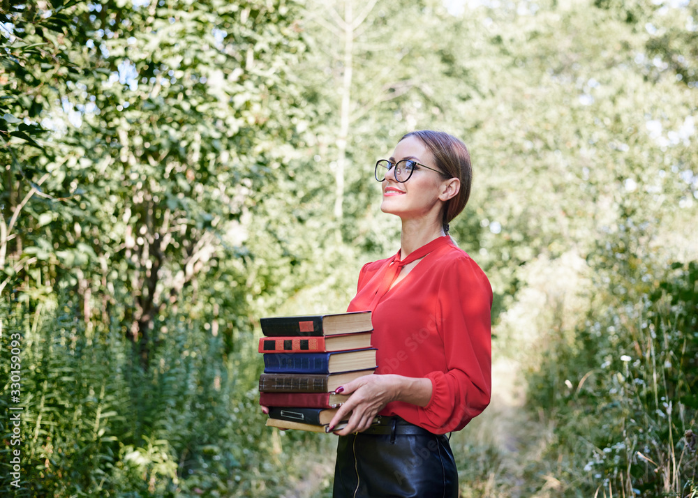 Fototapeta premium Three-quarter portrait of young woman, wearing eyeglasses with tied hairstyle, holding a bunch of books. Student with books in the park. Studying process. Book lover.Leisure time in campus.